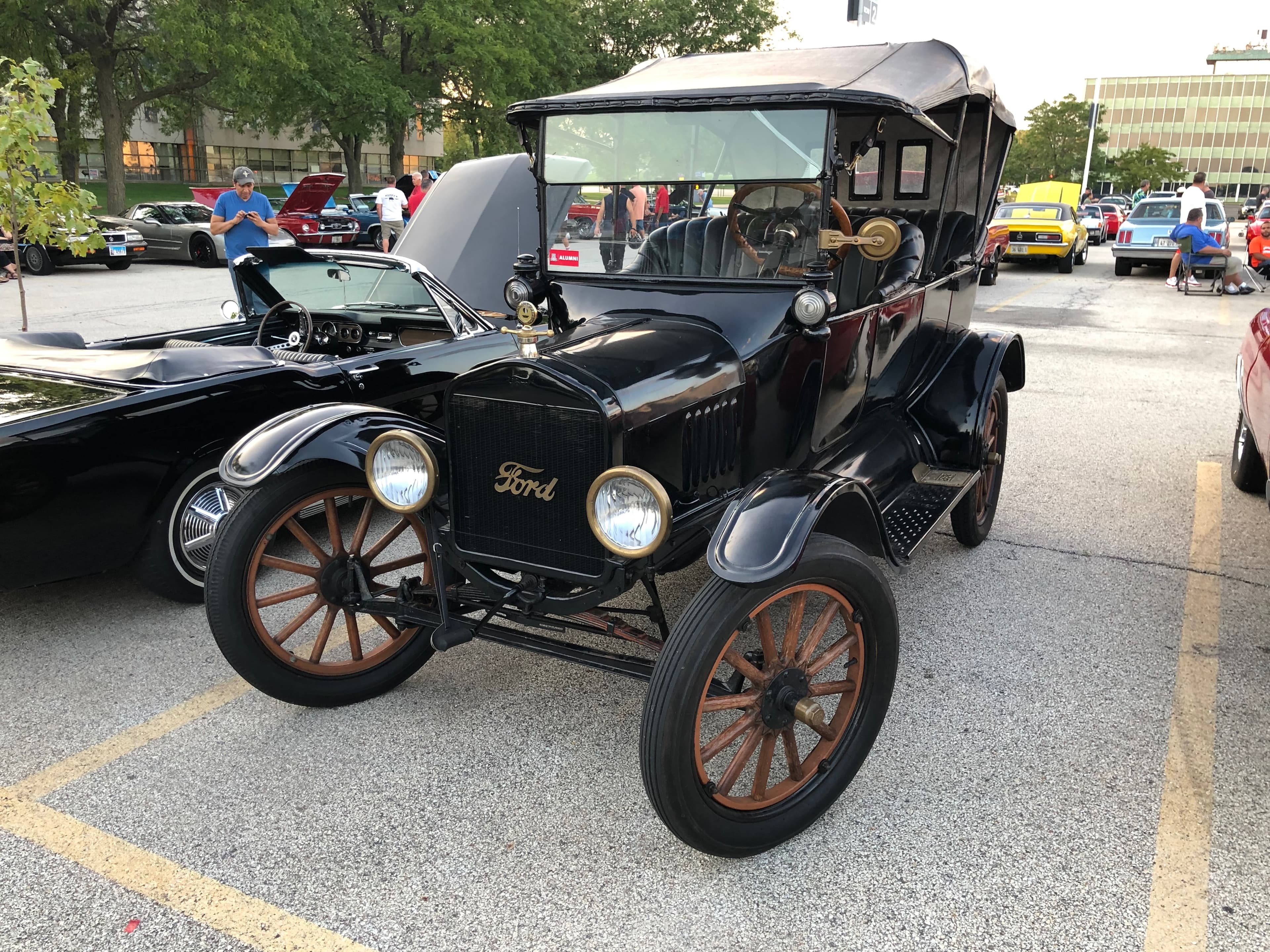 A black Ford Model T from the early 20th century, with wooden-spoke wheels and a convertible top, is parked at an outdoor car meet. It has a prominent front grille with the Ford logo, two round headlights, and is surrounded by other cars and people.