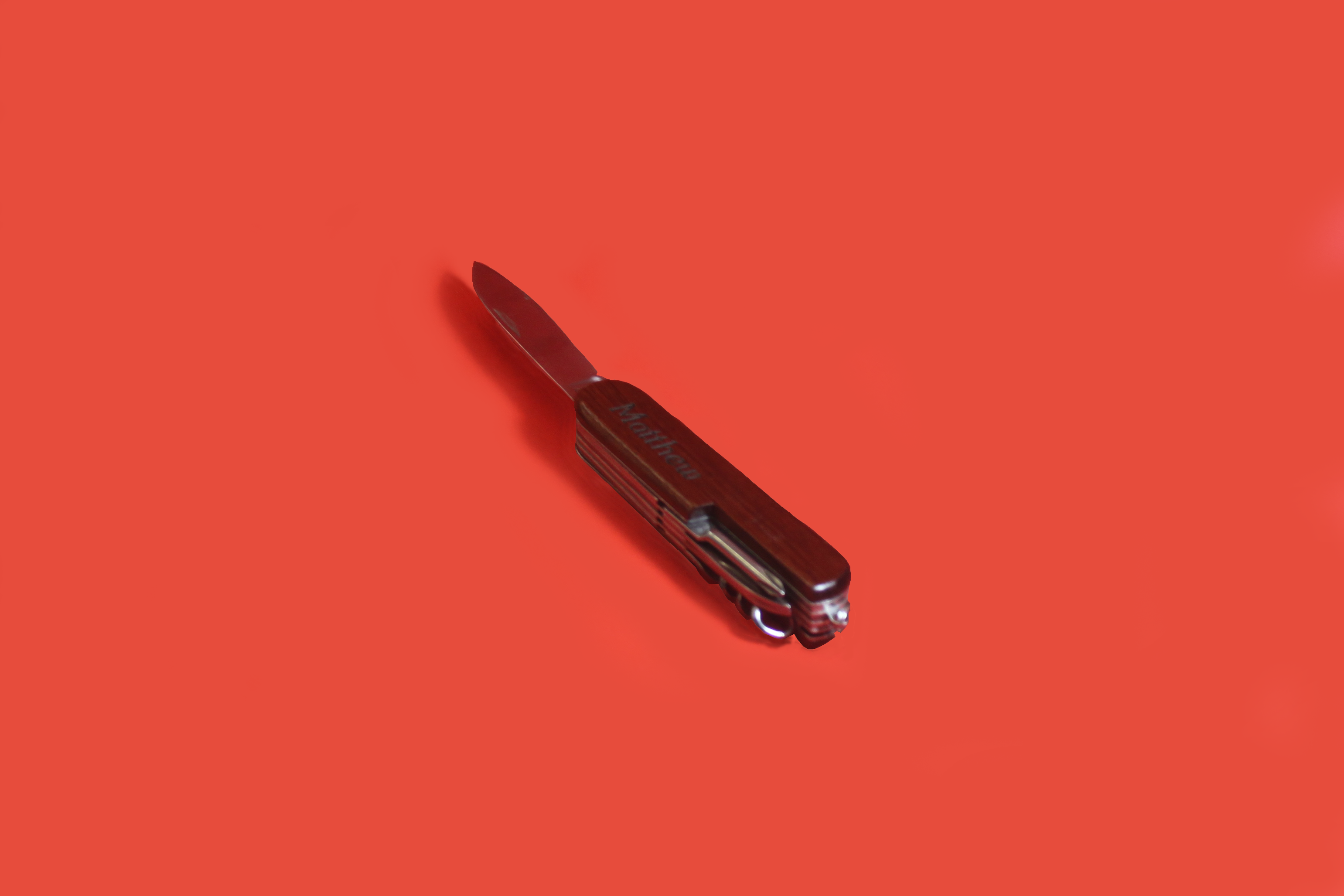A small switchblade is set on a red background, casting a modest shadow. The wooden handle has the name Matthew engraved into it.