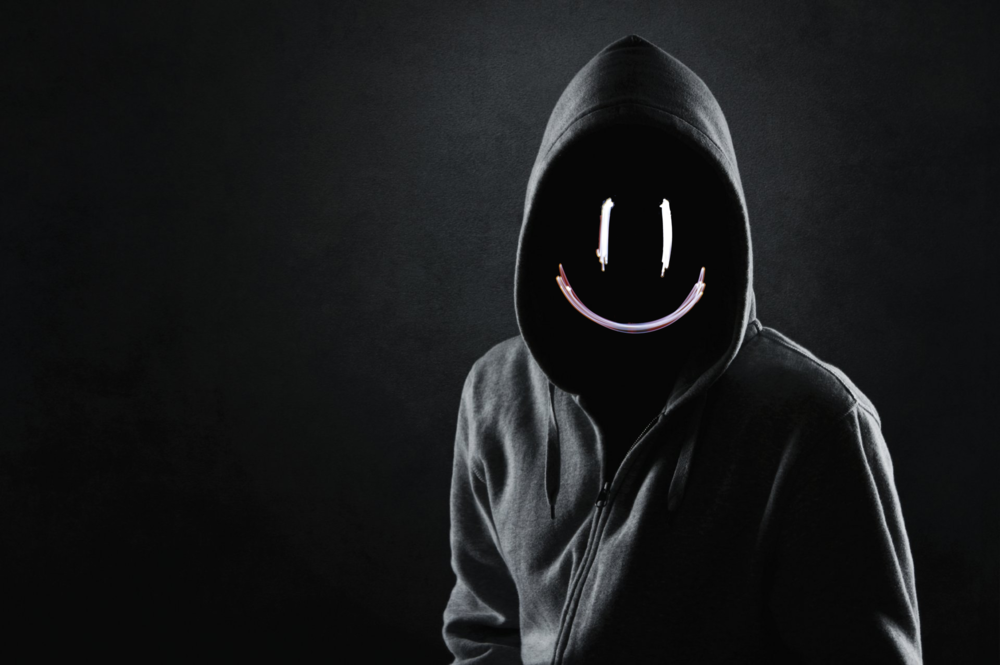 A hooded figure stands manacingly in the dark, with a florescent smiley face in place of a normal face.