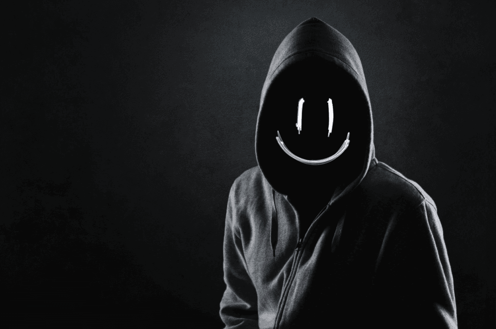 A hooded figure stands manacingly in the dark, with a florescent smiley face in place of a normal face.