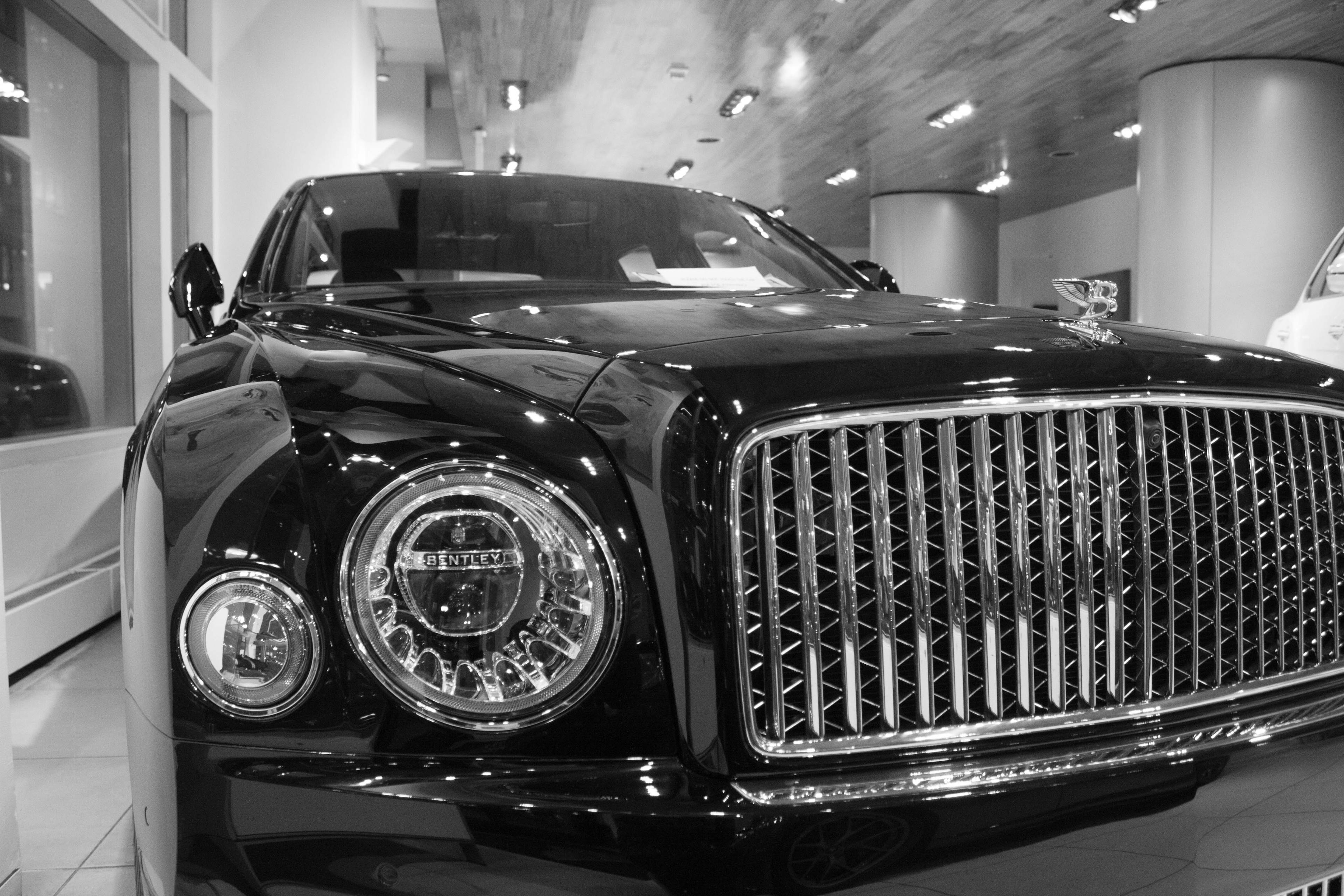 The front fascia of a black Bentley Mulsanne inside a showroom. The entire image is monochromatic and angled from a crouching position near the right headlight.